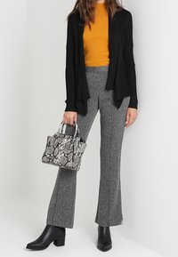 Kobieta w musztardowej bluzce, czarnym kardiganie, szarych spodniach, czarnych botkach za kostkę, trzymająca torebkę w wzór węża.