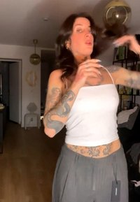 Žena s tmavými vlasmi a tetovaniami na rukách a bruchu, v bielom tielku a sivých nohaviciach, si upravuje vlasy v obývacej izbe.