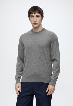 Giovane uomo che indossa un maglione grigio semplice a maniche lunghe e jeans blu scuro, in piedi contro uno sfondo chiaro e uniforme.