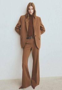 Bruin op maat gemaakt pak met een aansluitende jas, wijde broekspijpen en een bruine coltrui. Heeft een subtiele textuur en een zwarte riem met een ovale gesp.