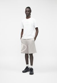 Balti trumpomis rankovėmis marškinėliai su įspaustu logotipu, derinami su smėlio spalvos šortais, ant kurių yra didelis šviesiai pilkas grafikas. Juodi sportbačiai ir balti kojinės.