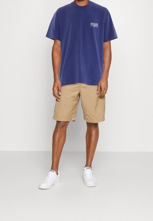 Blaues Baumwoll-T-Shirt mit dem Schriftzug "PEQUS" auf der Brust, kombiniert mit beigen Shorts. Das Modell trägt weiße Sneakers und steht vor einem grauen Hintergrund.