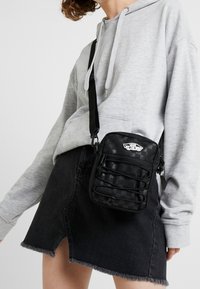 Schwarze gewebte Umhängetasche mit strukturiertem Oberflächen, verstellbarem Tragegurt und einem Reißverschlussfach. Mit einem Logo-Patch auf der Vorderseite.
