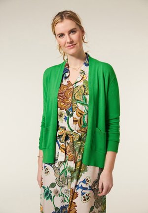 Groene cardigan van zacht materiaal, open voorkant en ribbelmanchetten. Gedragen over een jurk met bloemenpatroon in verschillende tinten groen, bruin en blauw.