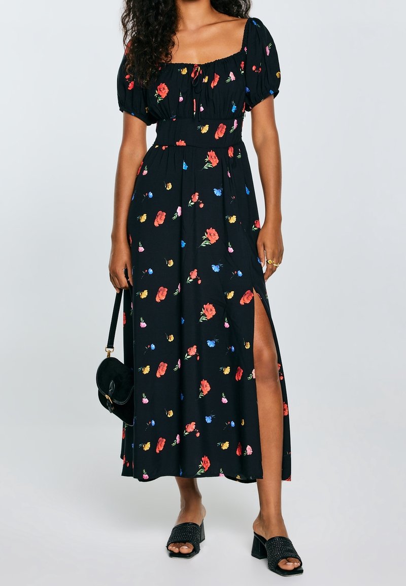 Femme portant une robe midi noire à motifs floraux avec des manches bouffantes et une fente à la cuisse, tenant un sac à main noir et portant des sandales noires à talons.