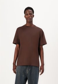 Braunes, kurzärmliges T-Shirt aus glattem Stoff, mit Rundhalsausschnitt und lockerer Passform. Getragen mit locker sitzenden blauen Jeans.