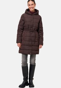 Bruine pufferjack met een getailleerde taille, capuchon en drukknoopsluiting. Getextureerde stof met zichtbare horizontale stiksels. Gecombineerd met grijze jeans en zwarte laarzen.