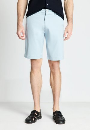 Uomo con pantaloncini azzurri fino al ginocchio, scarpe nere intrecciate in pelle e camicia nera su uno sfondo bianco semplice.