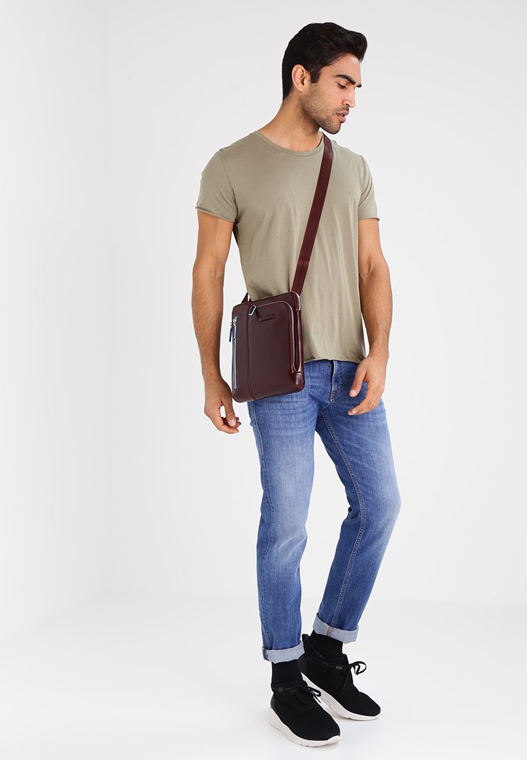 Borsa a spalla in pelle marrone con una tasca con zip, forma rettangolare, tracolla regolabile, indossata con jeans azzurri chiari e una maglietta grigia.
