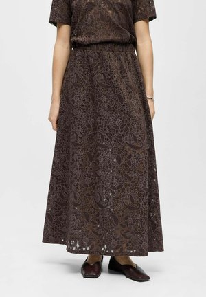 Femme portant une jupe longue marron à motif floral avec une taille élastique et des ballerines pointues marron foncé sur fond blanc.