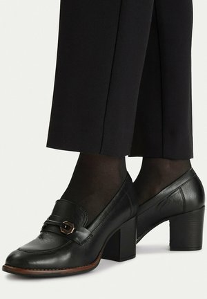 Schwarze Leder-Loafer mit Blockabsatz, getragen mit schwarzen transparenten Strumpfhosen und schwarzen, kurzen Hosen vor einfachem Hintergrund.