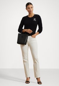 Svart kabelstrikket genser med sidelukking og logo, kombinert med lyse rett-lags jeans og en svart tote bag. Åpen-tå svarte sandaler.