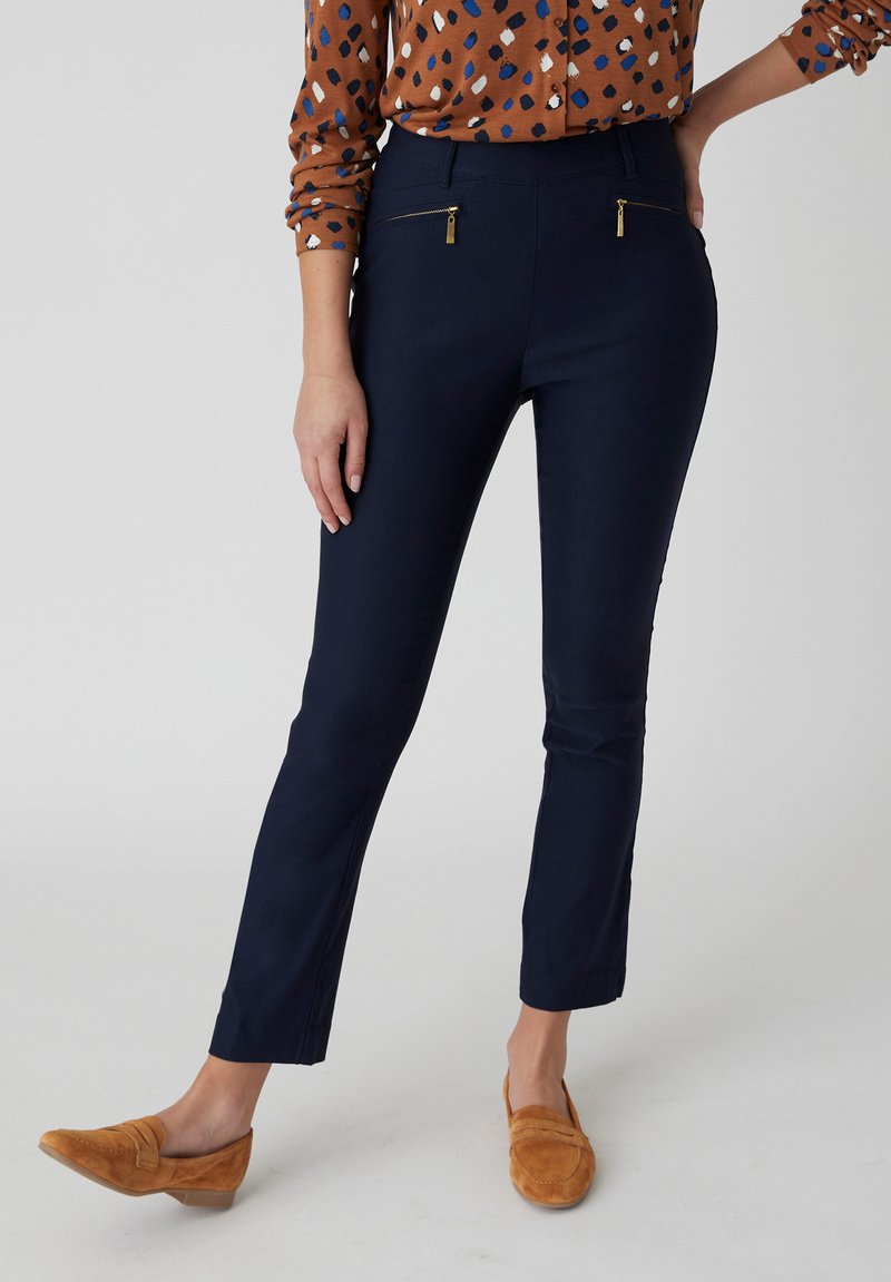 Pantaloni slim-fit blu navy con due tasche frontali con zip, realizzati in un tessuto morbido. Abbinati a una blusa marrone a fantasia e mocassini beige.