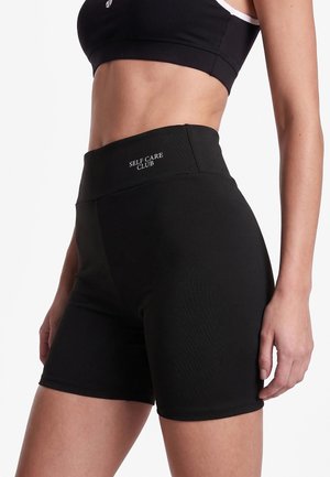Vrouw die zwarte, hoog getailleerde fietsshorts draagt met de tekst "SELF CARE CLUB" en een zwarte sportbeha tegen een effen witte achtergrond.