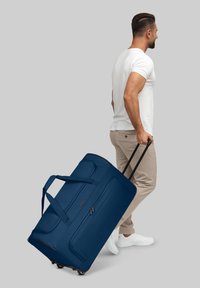 Marineblauwe rolduffelbag met een gestructureerde oppervlakte, meerdere vakken, versterkte handvatten en zwarte wielen, wordt getrokken door een persoon.