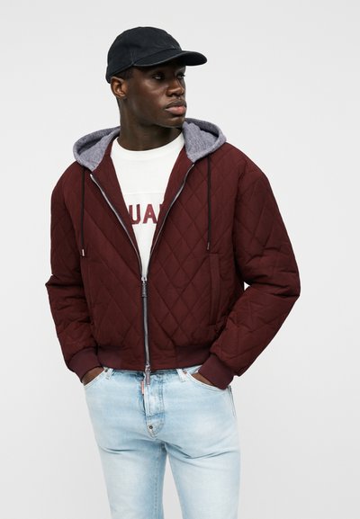 Homme portant une veste matelassée zippée de couleur rouge foncé avec une capuche grise, un t-shirt blanc à motif, un jean bleu clair et une casquette de baseball noire, les mains dans les poches.