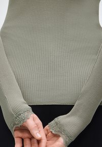 Χέρια ενωμένα πίσω από την πλάτη, φοράει πράσινη ριμπ μακρυμάνικη μπλούζα με δαντέλα στις μανσέτες και μαύρο παντελόνι.