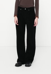 Pantaloni a gamba larga in corduroy nero con tasche frontali, chiusura con bottone in metallo e una finitura testurizzata. Abbinati a un top marrone a maniche lunghe.
