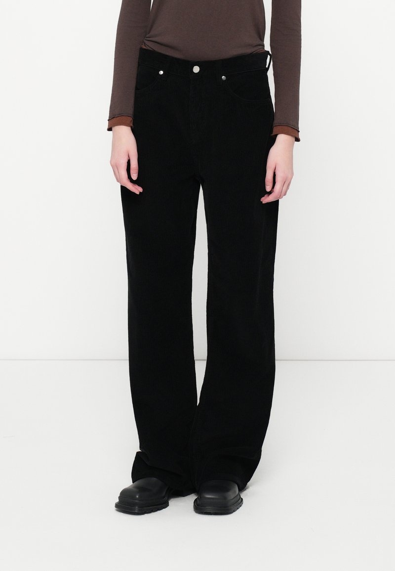 Pantaloni a gamba larga in corduroy nero con tasche frontali, chiusura con bottone in metallo e una finitura testurizzata. Abbinati a un top marrone a maniche lunghe.