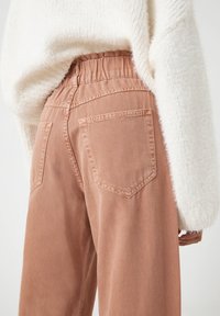 Kalhoty v broskvovém odstínu, volného střihu s elastickým pasem, obsahující dvě zadní kapsy a měkkou texturu látky. Nosené s bílým, huňatým svetr.