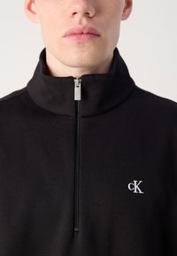 Čierne zapínacie mikiny na zips s vysokým golierom a diskrétnym logom cK. Má hladký povrch a kovový zips.