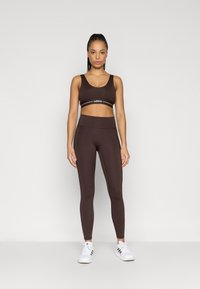 Sujetador deportivo marrón con banda blanca con logo, combinado con mallas marrones de talle alto. Zapatillas deportivas blancas completan el conjunto. Textura lisa y ajustada.