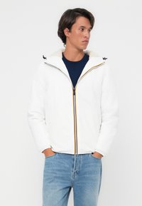 Hvid jakke med en tilsluttet hætte, der har et blødt indvendigt fôr og kontrastfarvede lynlåsdetaljer. Kombineret med blå jeans.