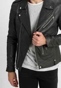 Veste de moto en cuir noir avec des zips et des pressions argentées, présentant une fermeture éclair frontale asymétrique, des épaulettes et des poches latérales.