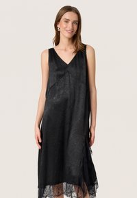 Schwarzes ärmelloses Kleid aus strukturiertem Stoff, mit V-Ausschnitt, asymmetrischem Saum und Spitzenakzenten entlang des unteren Randes.
