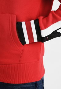 Persoon in een rode hoodie met hand in de voorzak; de mouw heeft een zwart, rood en wit gestreepte manchet met een klein logo detail.