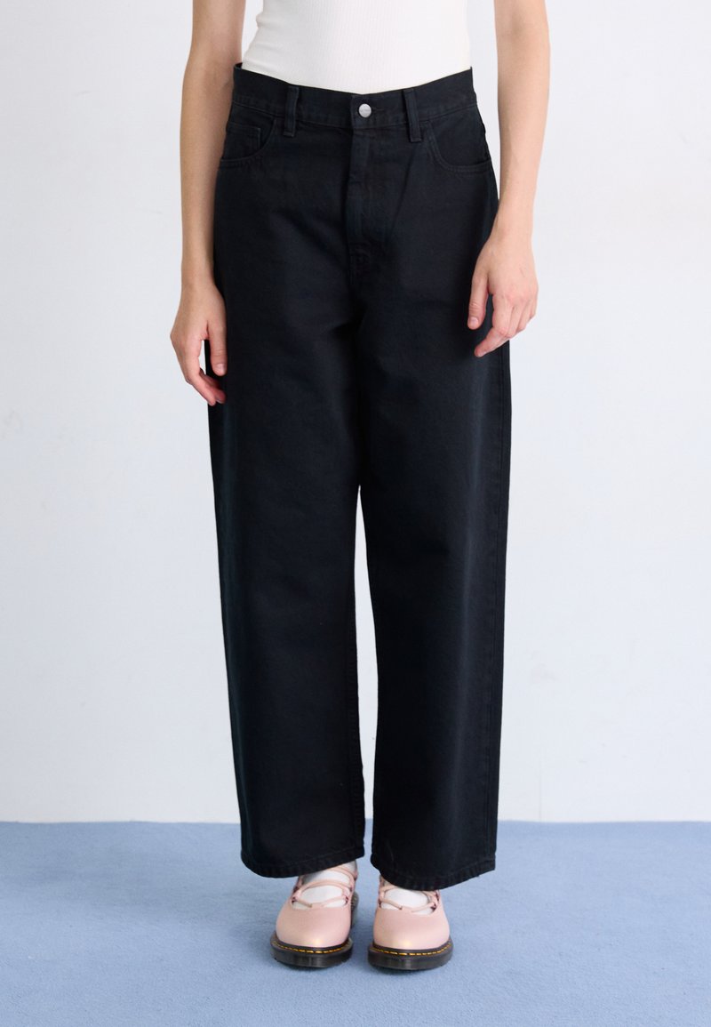 Osoba oblečená v čiernych voľných nohaviciach s vysokým pásom, ružových šnurovacích topánkach a bielych ponožkách, stojaca na svetlomodrej podlahe na pozadí bielej steny.