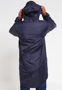 Laivastonsininen vedenpitävä pitkä takki hupulla, jossa on vetoketjutasku takana ja sileä, kevyt materiaali. Sisältää pitkät hihat.