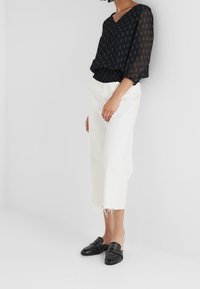 Svart genomskinlig blus med V-ringning och mönstrad textur, i kombination med vita urklippta byxor och svarta slip-on skor med nitar.