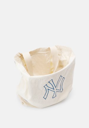 Kermansävyinen kangaskassi, jossa on kaksi kahvaa ja sininen tyylitelty "NY"-logo edessä yksivärisellä taustalla.