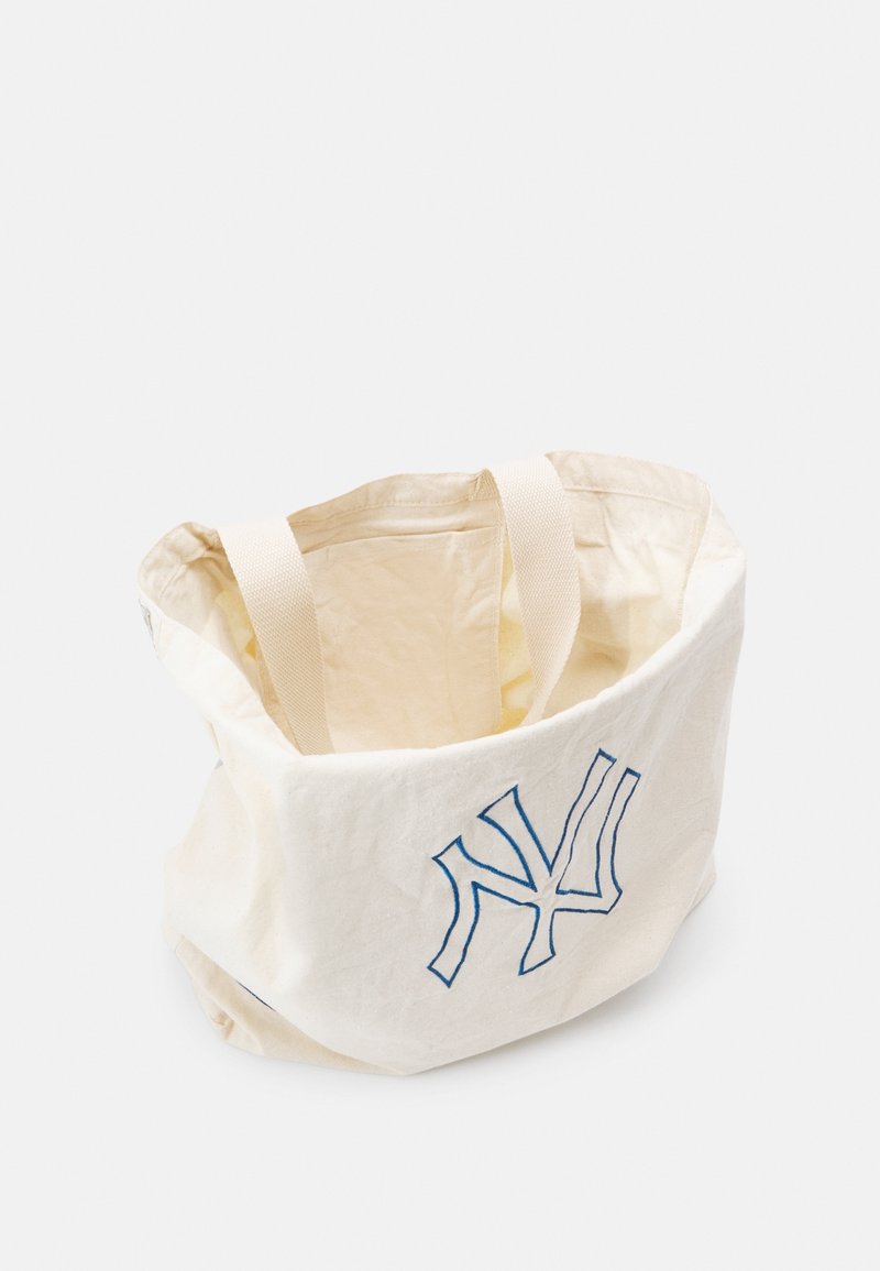 Sac cabas en tissu couleur crème avec deux anses et un logo «NY» stylisé en bleu sur le devant, sur un fond uni.