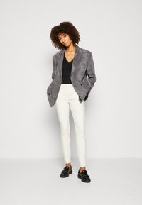 Blazer en suède gris sur un haut noir, associé à un pantalon slim blanc et des mocassins noirs avec des franges, sur un fond uni.