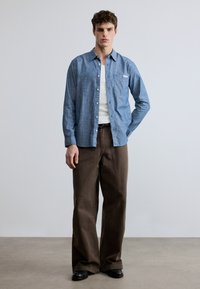 Svetlo modra srajca z dolgimi rokavi in gumbi, kombinirana z ohlapnimi rjavimi hlačami. Srajca ima prsni žep in teksturirano tkanino.