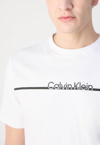 Hvid bomulds t-shirt med rund hals, prydet med en sort vandret linje og "Calvin Klein" trykt med fede sorte bogstaver.