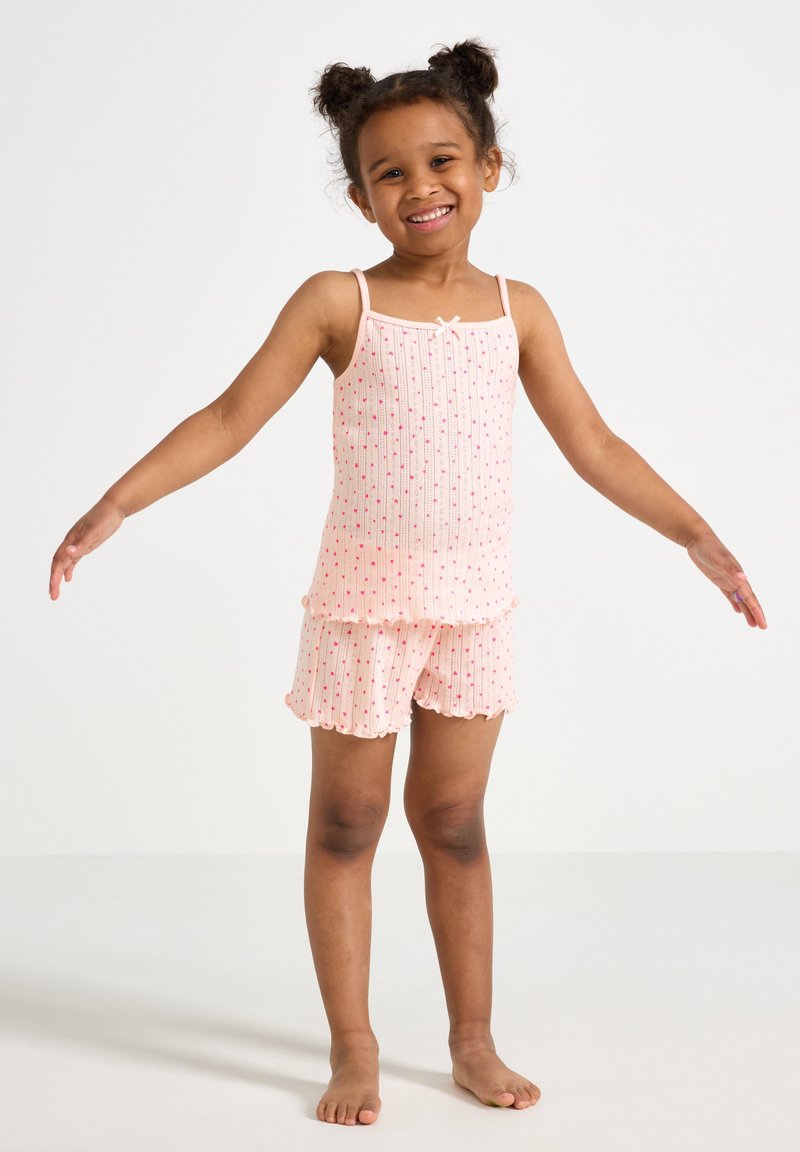 Valkoinen puuvillainen yöasu, jossa on ribbitekniikalla valmistettu toppi ohuilla olkaimilla ja liehukehelmalla, yhdistettynä vastaaviin shortseihin, joita koristavat pienet punaiset yksityiskohdat.