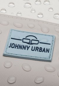 Vízálló táska felülete vízcseppekkel. Kék gumilogo címke "JOHNNY URBAN" felirattal, texturált, világos színű háttérrel.