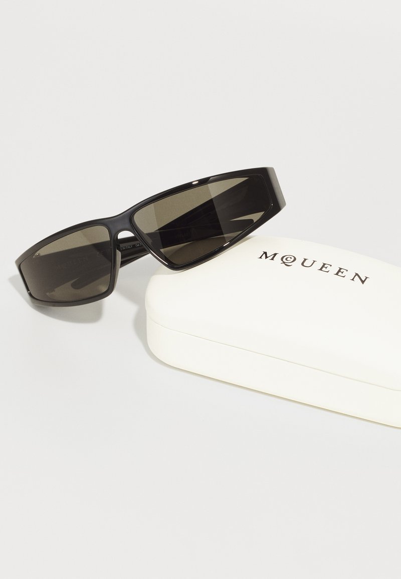 Occhiali da sole neri con lenti rettangolari, finitura opaca e design elegante, posizionati accanto a un astuccio bianco con la scritta "MQUEEN" stampata in nero.