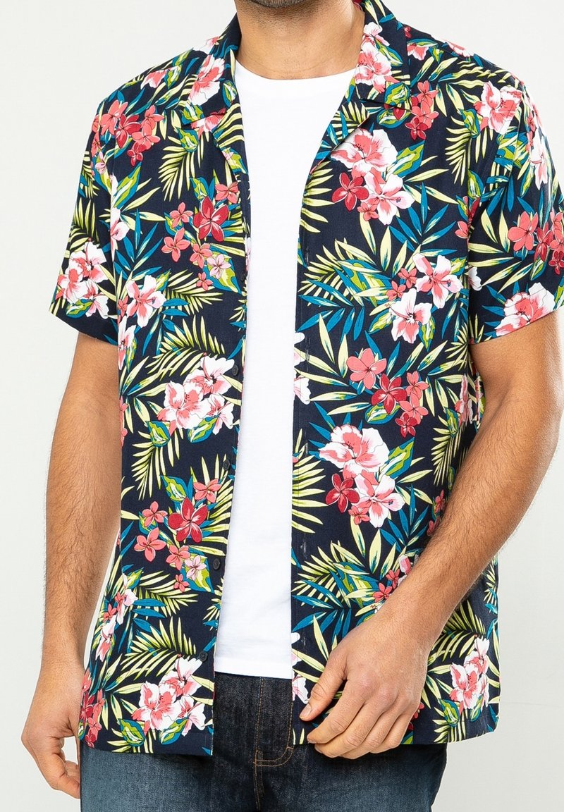 Hombre vistiendo una camisa de manga corta azul marino con estampados florales en rojo, rosa y blanco sobre una camiseta blanca básica y jeans azules.