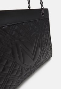 Czarna pikowana skórzana torebka z łańcuszkowymi uchwytami, z dużym wyklejonym logo na przodzie i gładkim skórzanym wykończeniem u góry.