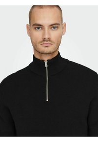 Suéter negro de cuello alto acanalado con una cremallera plateada en el frente, hecho de un tejido texturizado y con una silueta ajustada.