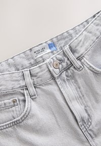 Jean en denim gris clair à jambe large, taille haute, bouton en métal, passants de ceinture et étiquette indiquant la taille et l'origine.