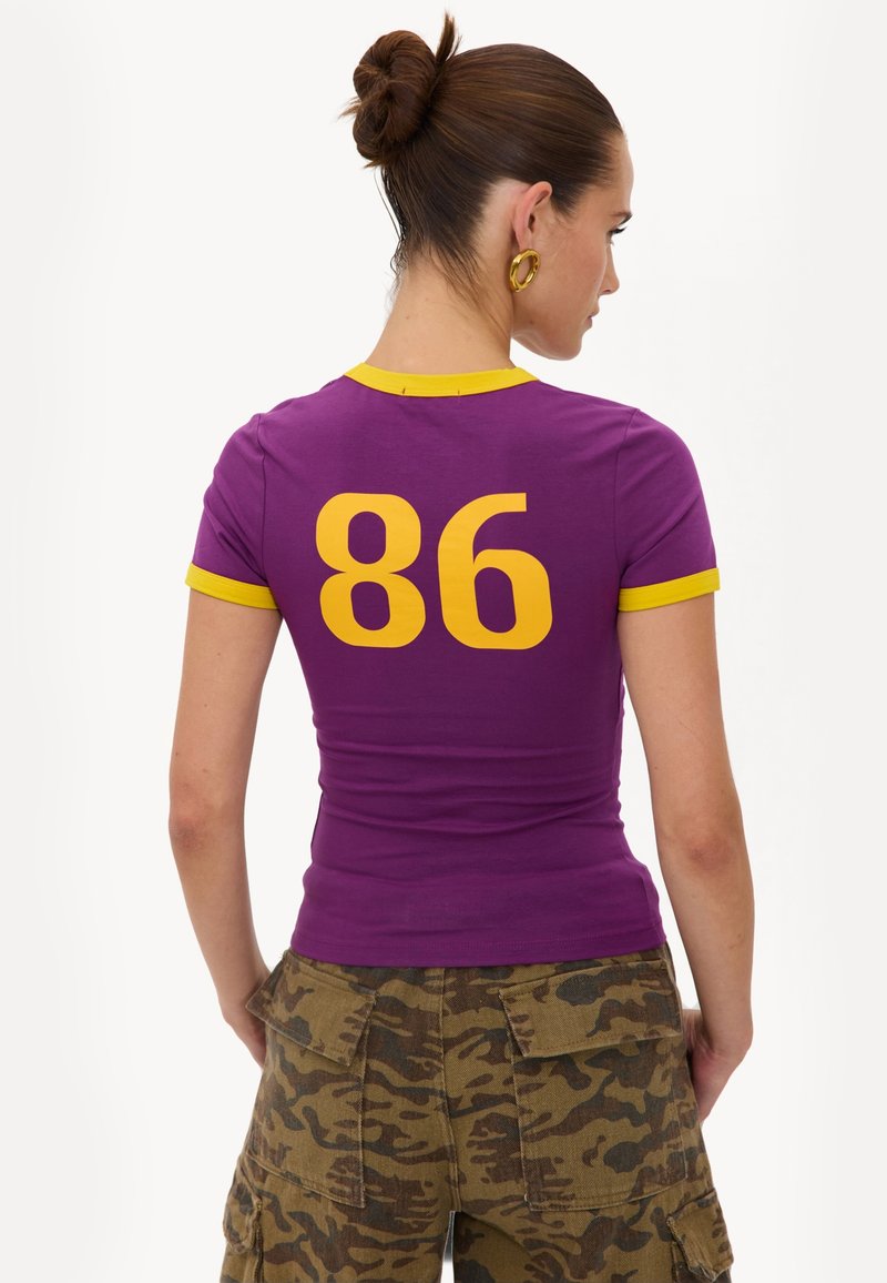 Femme aux cheveux en chignon portant un t-shirt ajusté violet avec le numéro 86 jaune dans le dos et une bordure jaune, associé à un pantalon cargo camouflage.