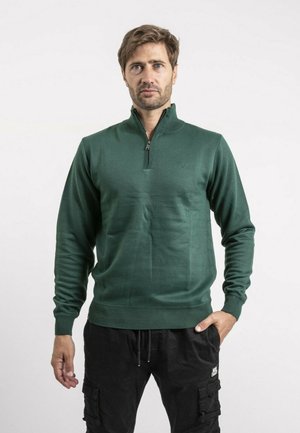 Homme aux cheveux châtain clair portant un pull vert foncé à fermeture éclair jusqu'au quart et un pantalon cargo noir, debout avec une main dans la poche.