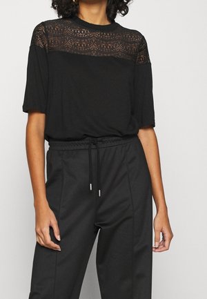 Kobieta ubrana w czarną bluzkę z krótkim rękawem i koronkowym karczkiem oraz czarne spodnie ze sznurkiem, stoi na tle jednolitego jasnego tła.