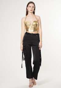 Gouden metalen cropped top met spiraalontwerp, gecombineerd met zwarte cargobroek met zijzakken en strikjes. Model draagt gouden spannerhakken.