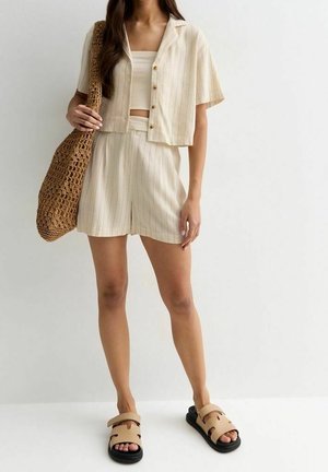 Femme portant un short beige à fines rayures et une chemise à manches courtes assortie avec boutons, des sandales beiges, et portant un grand sac cabas marron en crochet.